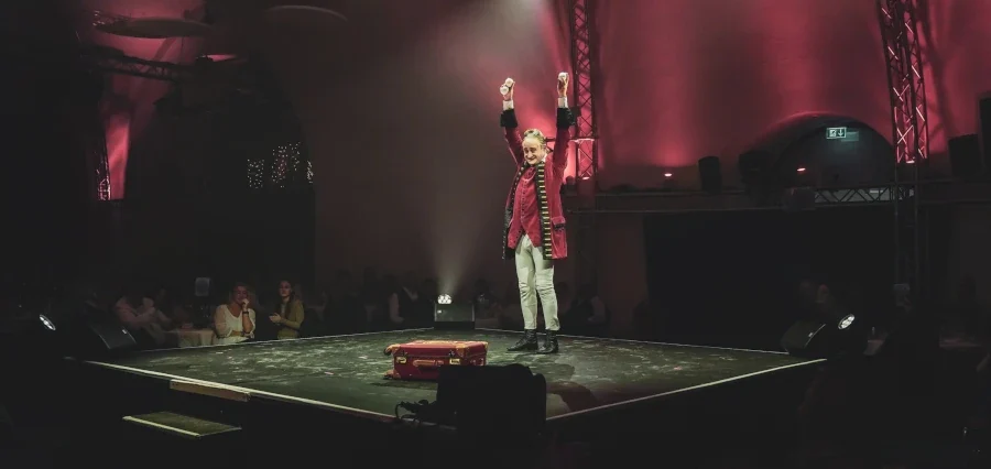 Stephan ;asur Le Comte Jongleur & barocker Zeremonienmeister Ein Schauspieler auf der Bühne, umgeben von Publikum, mit roten Lichtern im Hintergrund.