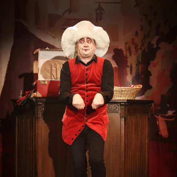 Stephan Masur komische Hutnumer Chapeaugraphie Clown in rotem Outfit mit weißem Hut steht vor einem Tisch auf einer Bühne.